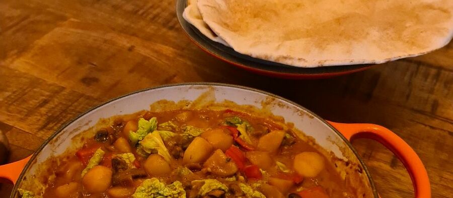 Eine Schale voll Curry mit orientalischem Löffel steht auf einem Holztisch vor einem Teller mit Fladenbrot.