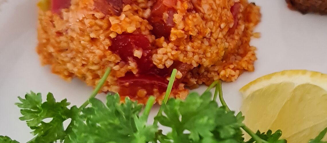 Ein weißer Teller auf dem neben Bulgursalat, eine Zitronenscheibe und grünen Salatblätter liegen.